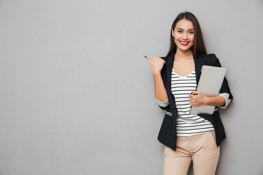 Smiling Asian Business Woman Holding Laptop Computer And Pointing Away