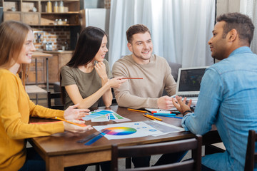 Interesting. Calm cheerful young man holding a convenient modern laptop while sitting at the table with his creative clever colleagues and discussing their strategy together