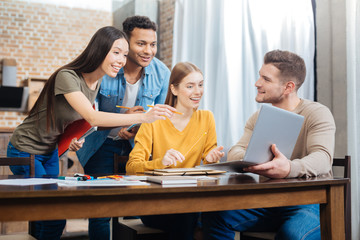 Amazing. Young smiling student holding a modern laptop while his positive friends looking attentively at the screen of it and feeling surprised