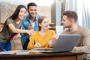 Great idea. Positive emotional students smiling and feeling excited while pointing at the screen of a modern laptop and generating marvelous ideas connected with their new project