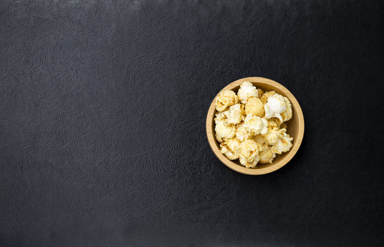 Salted popcorn in round wooden bowl on black leather texture background, movie night snack