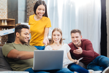 Obraz na płótnie Canvas Marvelous. Happy emotional young friends sitting comfortably on a big soft sofa and smiling cheerfully while looking at the screen of a modern laptop