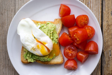 Perfect poached egg on a toast with smashed avocado and fresh tomatoes on a side on  a white plate on the wooden table