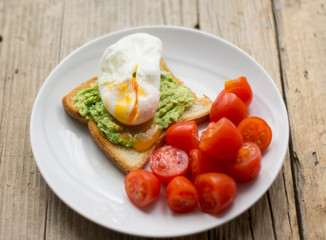 Perfect poached egg on a toast with smashed avocado and fresh tomatoes on a side on  a white plate on the wooden table