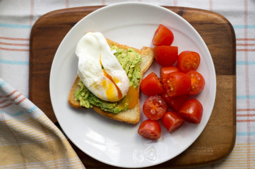 Perfect poached egg on a toast with avocado with tomatoes on a side on a white plate on a wooden cutting board