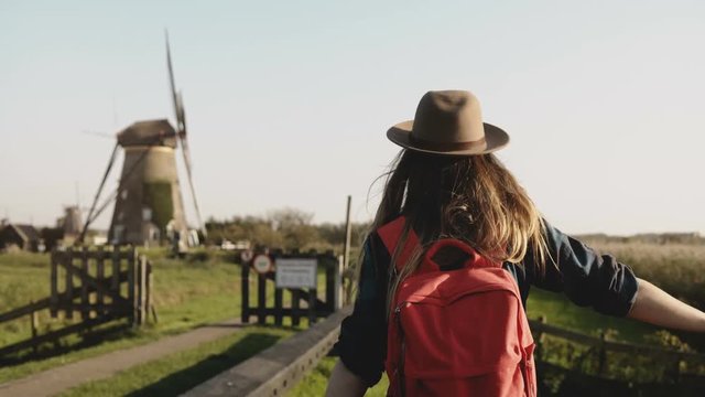 Local girl slowly walks up to a windmill farm. Woman in hat with long hair and red backpack coming back home. 4K.
