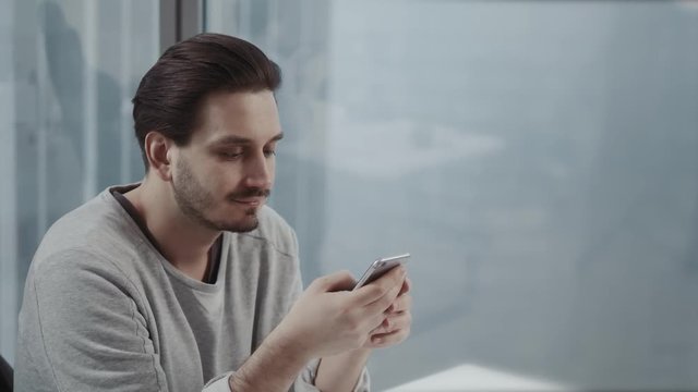 Portrait of young businessman talk on cellphone while stand by his office window in modern interior of skyscraper building, male entrepreneur having mobile phone conversation after important briefing