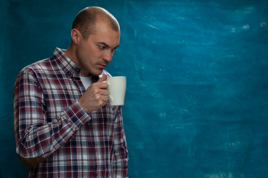 Serious man wearing plaid shirt posing with coffee mug in hands against blue background