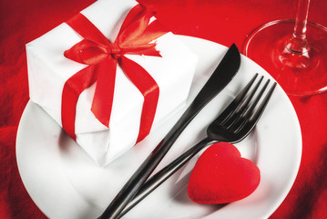 Valentine's day table setting with plate, fork, knife, gift box and red heart, on red tablecloth background copy space