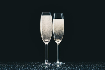 two glasses with champagne with drops on table on black