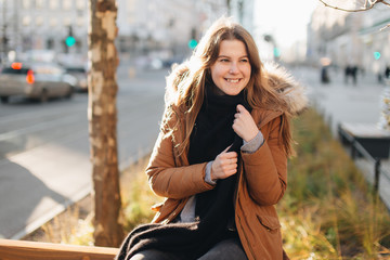 Young pretty girl sit on the bench. Hang her scarf in one hand and look around. Winter clothing with warm weather sunset scene