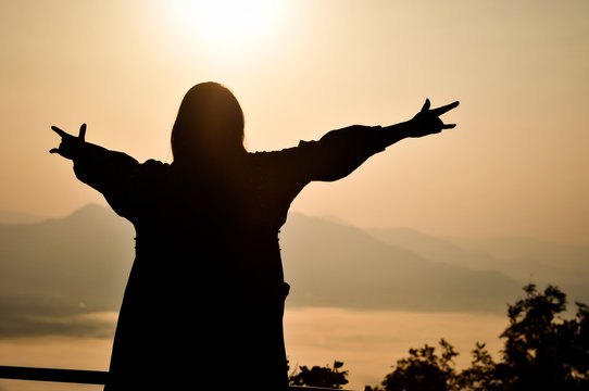 Silhouette of woman put her hand with 
Love symbol up in the air with background of early morning foggy sunrise on top of mountain soft focus.