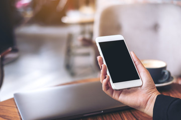 Mockup image of a woman's hand holding white mobile phone with blank black desktop screen and laptop on wooden table in cafe