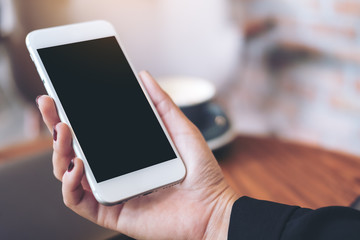 Mockup image of a woman's hand holding white mobile phone with blank black desktop screen and laptop on wooden table in cafe