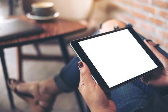Mockup top view image of a woman wearing ripped jean sitting cross legged , holding black tablet pc with blank white desktop screen on thigh in cafe