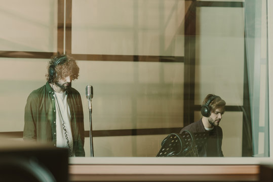 band of two musicians recoding song behind glass at studio