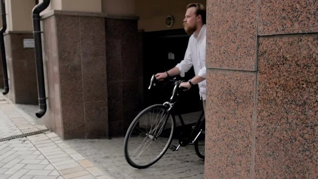 Slow motion video of handsome bearded man walking out of passageway and getting on vintage sport bicycle