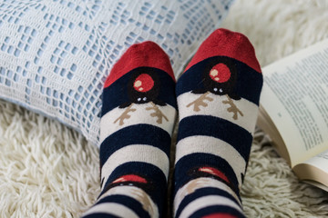 Close-up of female legs stretched over white carpet in Christmas socks and open book, shallow depth of focus. Concept, winter lazy days.