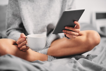 Woman drinking coffee and holding tablet while sitting on the bed in the morning.