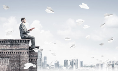 Man on roof edge reading book and cityscape at background