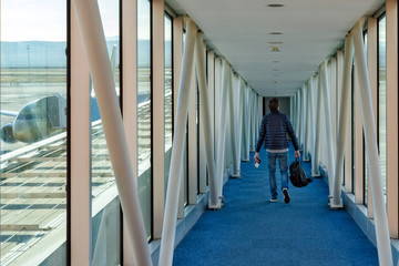 A man with a backpack in his hands walks down the corridor to board the plane. Out the window feed part of the aircraft. - Powered by Adobe