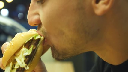 Close up of young man eating hamburger or cheeseburger indoor at evening. Side view of male mouth biting and chewing burger. Slow motion