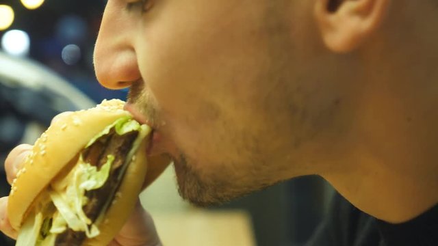 Close up of young man eating hamburger or cheeseburger indoor at evening. Side view of male mouth biting and chewing burger. Slow motion