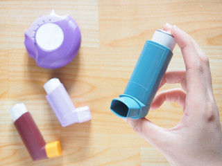 Patient's hands holding blue asthma inhaler with set of asthma/COPD inhalers on wood table....