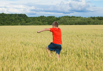 Fototapeta na wymiar The boy in a bright T-shirt runs along the yellow field where ears of grain grow, the grain against the blue sky, the rear view.