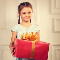 Beautiful cute kid girl holding big present box with red ribbon bow and looking happy on it in fashion blue dress on studio background. Toned vintage portrait with empty copy space