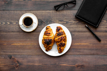 Work at breakfast. Coffee and croissants near notebook and glasses. Dark wooden background top view copy space