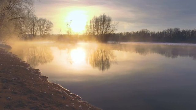 A winter walk along the shore of a hot spring flowing into the river. Frosty morning, beautiful winter landscape with a frozen river, soft light of the rising sun. Nice reflections on water surface.