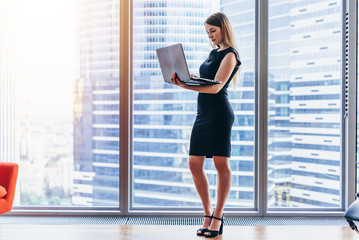 Businesswoman holding laptop standing in modern office against window with city view