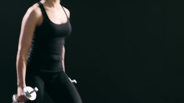 A wonderful view of a sportive woman in a black suit who does forward lunges while keeping metallic dumbbells in her hands in a black studio 