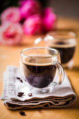 Valentine`s day celebration concept. Two cups of coffee with froth on top on beige napkin in double bottom glasses with heart love shape sugar on wooden table background with pink flowers