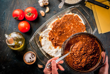 Woman cooking homemade classic lasagna bolognese, on dark blue table; with ingredients, top view copy space, hands in picture
