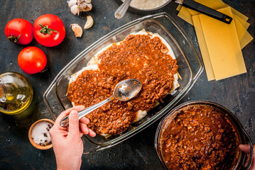Woman cooking homemade classic lasagna bolognese, on dark blue table; with ingredients, top view...