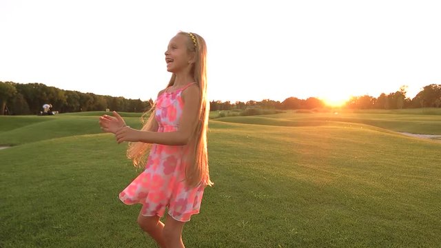 Happy child running over green meadow. Slow motion joyful little girl in cute dress having fun on beautiful nature background, sunset sky. Childhood and weekend in countryside.