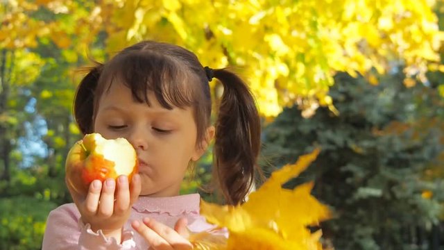 A beautiful little girl in an autumn park eats an apple.