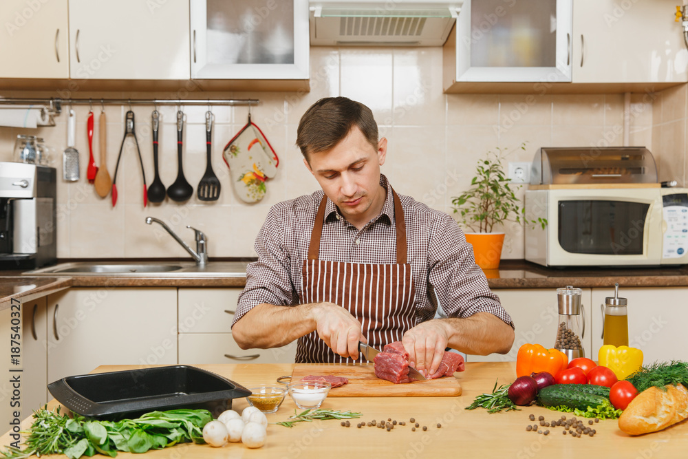 Wall mural Attractive caucasian young man in apron sitting at table with vegetables, cooking at home preparing meat stake from pork, beef or lamb, in light kitchen with wooden surface, full of fancy kitchenware.