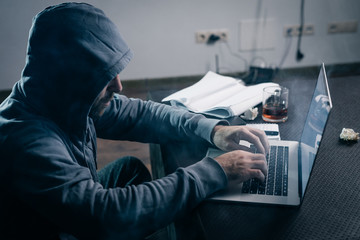 young guy hacker in a sweatshirt with a hood on his head sits behind a laptop