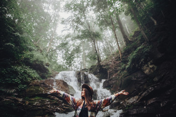 happy stylish traveler girl in hat relaxing at waterfall, atmospheric moment of success. hipster woman with backpack travelling, hands up. space for text. wanderlust and travel concept