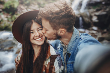 happy hipster couple taking selfie at waterfall in forest. stylish family traveling, embracing and...
