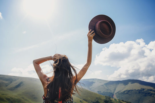 stylish traveler woman looking at mountains. hipster girl on top of mountain, raising hands and holding hat in sky. space for text. atmospheric epic moment. wanderlust and travel concept