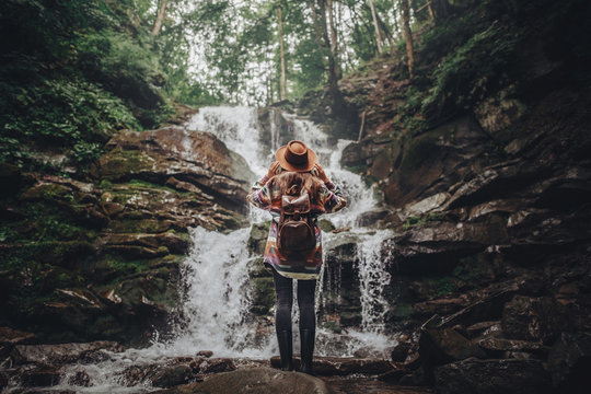 wanderlust and travel concept. stylish traveler girl holding hat looking at waterfall, exploring woods. hipster woman with backpack travelling at river in forest, moment. space for text