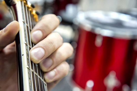 The guitarist's hand, close-up and soft focus, takes the akrod on a guitar fretboard, against the background of the drum set.