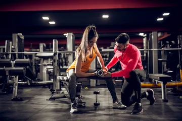 Foto op Aluminium Young healthy motivated attractive sporty flexible shape girl with a ponytail doing biceps exercises with dumbbells on the bench while handsome helpful personal trainer. © dusanpetkovic1
