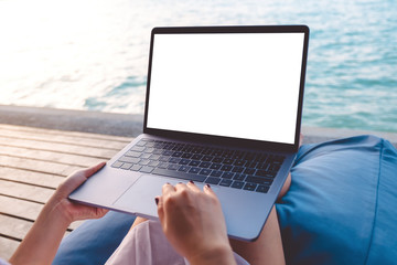 Mockup image of a woman using laptop with blank white desktop screen while sitting by the sea with blue sky background