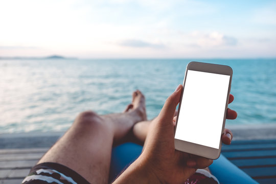 Mockup image of a man's hand holding white mobile phone with blank desktop screen sitting by the sea and blue sky background