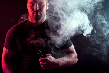 A young stylish man smoker in a black T-shirt is holding a    vape in his hand, and around and a big cloud of smoke from vape on a dark isolated background